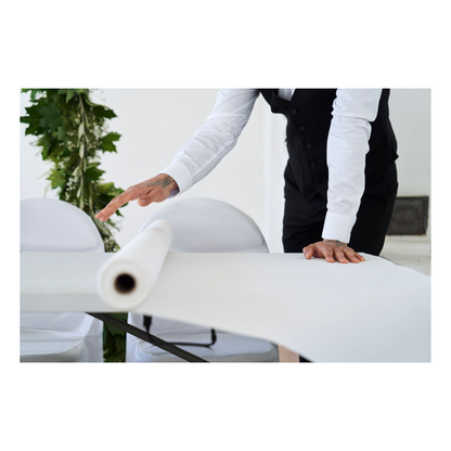 A person in evening attire, with a white shirt and black vest, rolls out a Dunicel tablecloth roll (1.18 m x 40 m) from Duni GmbH over a table; some green is visible against the plain background.