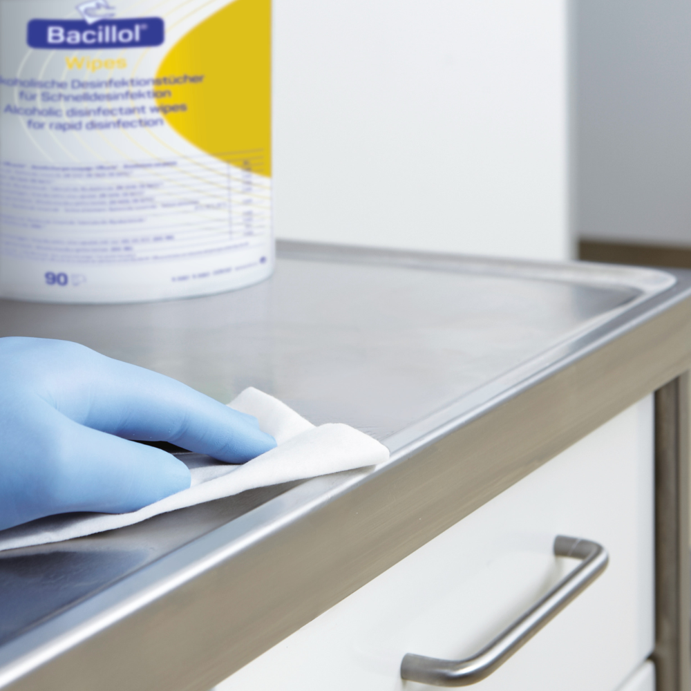 A gloved hand cleans a metal surface with a Paul Hartmann AG Hartmann Bacillol® Wipes disinfectant wipe, stand-up pouch, 90 wipes | piece (1 piece). The container with the Bacillol wipes is partially visible in the background and shows some text and design elements on the label. The metal surface appears to be part of a piece of furniture with a drawer.