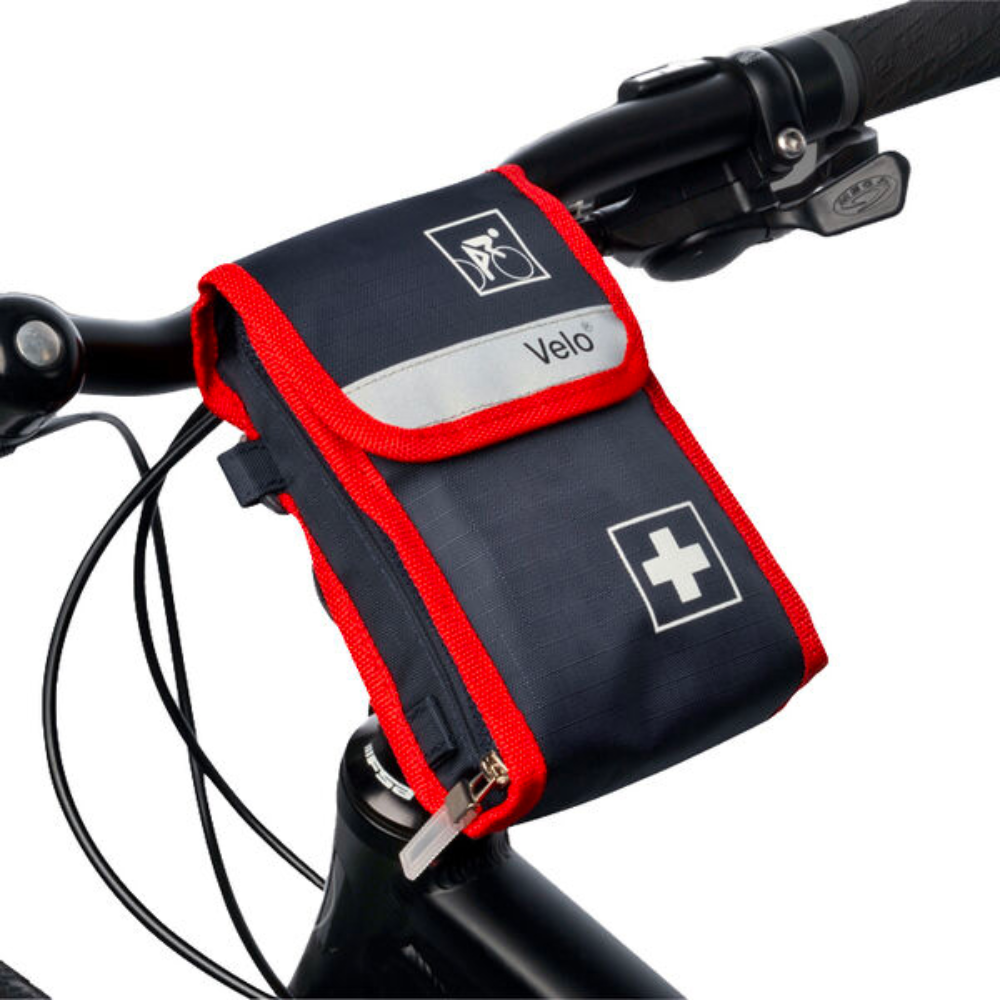 A small, black-red first aid kit with the inscription "Holthaus Velo® Bicycle First Aid Kit - 21-piece | Package (1 piece)" from Holthaus Medical GmbH & Co. KG is attached to the handlebar of a bicycle. The bicycle's first aid kit is marked with a white cross symbol and a bicycle symbol. Also visible are the handlebar and a part of the bicycle frame.