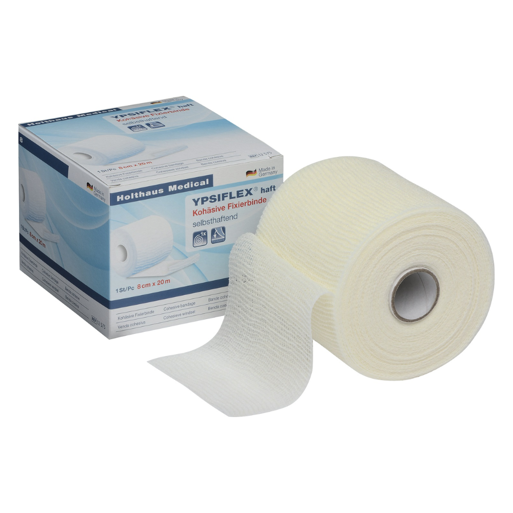 An open box with a roll of Holthaus Ypsiflex® adhesive elastic gauze bandage, 20 m. The roll is partially unrolled, revealing the textured white bandage. The box is labeled with product details and medical symbols from Holthaus Medical GmbH & Co. KG.