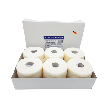 A box with six rolls of white medical adhesive tape labeled "Holthaus Ypsiflex® adhesive elastic gauze bandage, 20 meters" shows a small German flag symbol alongside other text. Each roll of Holthaus Ypsiflex® tape has a hole in the center, similar to elastic gauze bandage.