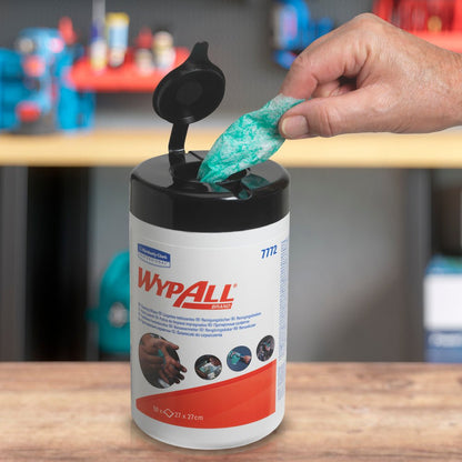 A hand pulls a green WypAll® wipe with high absorbency from an open tub of Kimberly-Clark GmbH (6 tubs x 50 sheets). Shelves with various items can be seen in the blurred background.