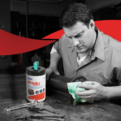A man in a work shirt wipes a metal object with a green WypAll® wipe from Kimberly-Clark GmbH in a workshop. On the table are the WypAll® wipes - Green (6 Tubs x 50 Sheets | Carton) and hand tools. The background is predominantly black and white with red accents.