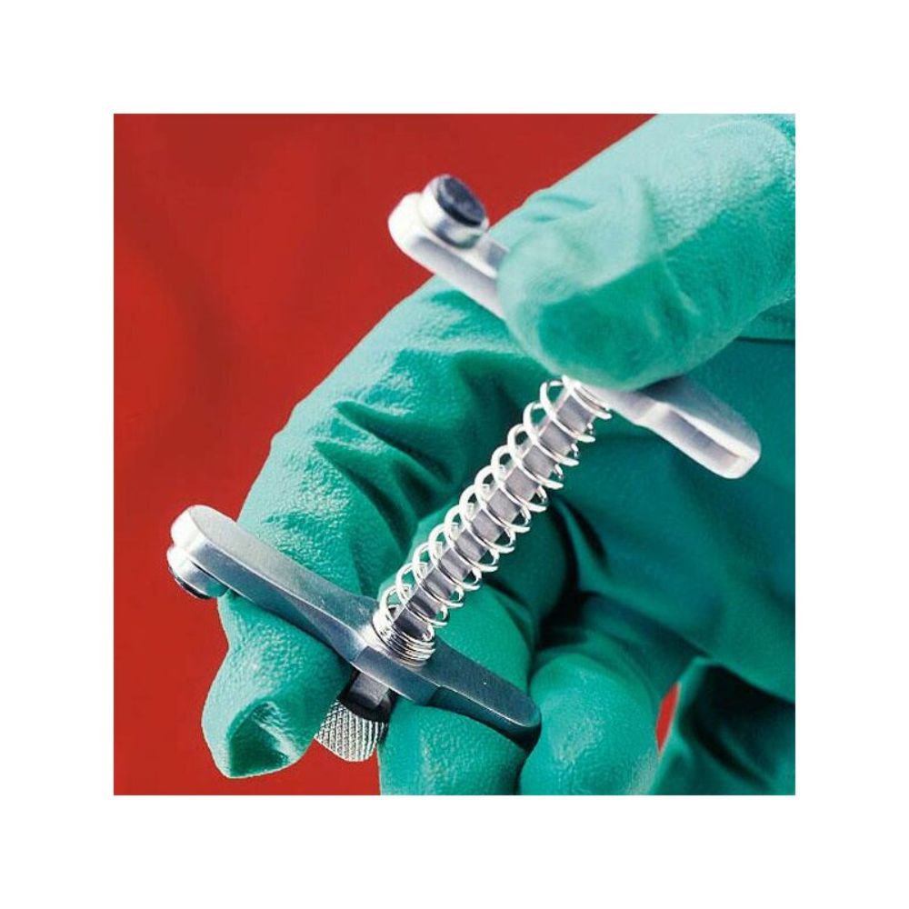 A person wearing green gloves holds an Altruan Servoprax mouth gag for cats with a cylindrical handle and screw thread against a red background, commonly used by veterinarians for veterinary procedures.