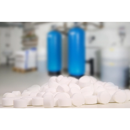 In the foreground, a stack of Regenit salt tablets according to EN 973 Type from K+S Minerals and Agriculture GmbH stands out, while the blurred background reveals two large blue water softening systems. The industrial setting suggests a precise utility space that complies with EN 973 Type A standards.