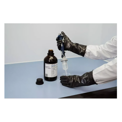 A person wearing AMPri SolidSafety ChemV Full Stable Brom-Butyl Chemical Protection Gloves with Viton outer layer and a white lab coat is using a pipette to draw liquid from a brown bottle on a light gray surface. The cap is nearby in the laboratory workspace, ensuring chemical resistance when handling substances.