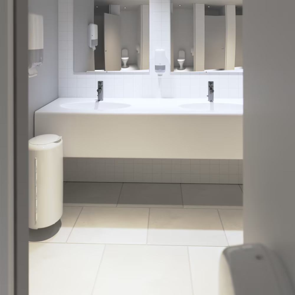 Eine öffentliche Toilette mit modernem, minimalistischem Design verfügt über einen Doppelwaschtisch mit Spiegeln darüber, TORK Tork 553000 Spender für Zickzack-Handtücher Elevation H3 | Packung (1 Stück) an der Wand und einen Abfalleimer auf dem Boden. Der Bereich ist weiß gefliest und sauber und gut beleuchtet.