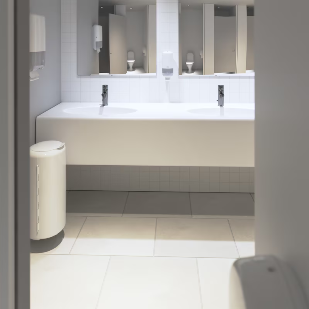 A clean, modern public restroom is seen with a long white counter with two sinks and faucets. Below the counter, a striking white tiled wall is noticeable. The room is equipped with mirrors, Tork soap dispensers from Essity Professional Hygiene Germany GmbH, and a white trash can on a tiled floor designed for high-traffic toilets.