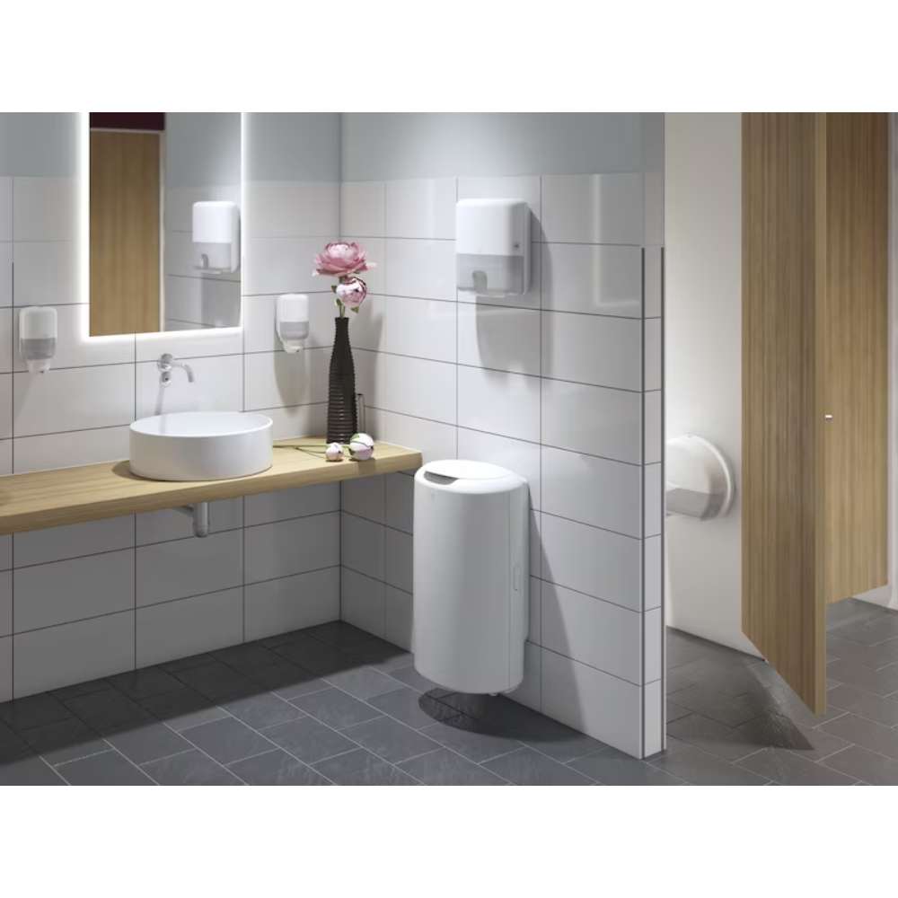 A modern public restroom with white-tiled walls and dark gray floor is a highlight of the Elevation design. It features a wooden shelf with a round washbasin, large mirror, Tork 555000 dispenser for Mini Jumbo Toilet Paper Elevation T2 from Essity Professional Hygiene Germany GmbH, as well as a soap dispenser, towel dispenser, flower vase, waste bin, and a partially open cubicle door.