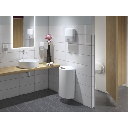 A modern public restroom with white-tiled walls and dark gray floor is a highlight of the Elevation design. It features a wooden shelf with a round washbasin, large mirror, Tork 555000 dispenser for Mini Jumbo Toilet Paper Elevation T2 from Essity Professional Hygiene Germany GmbH, as well as a soap dispenser, towel dispenser, flower vase, waste bin, and a partially open cubicle door.