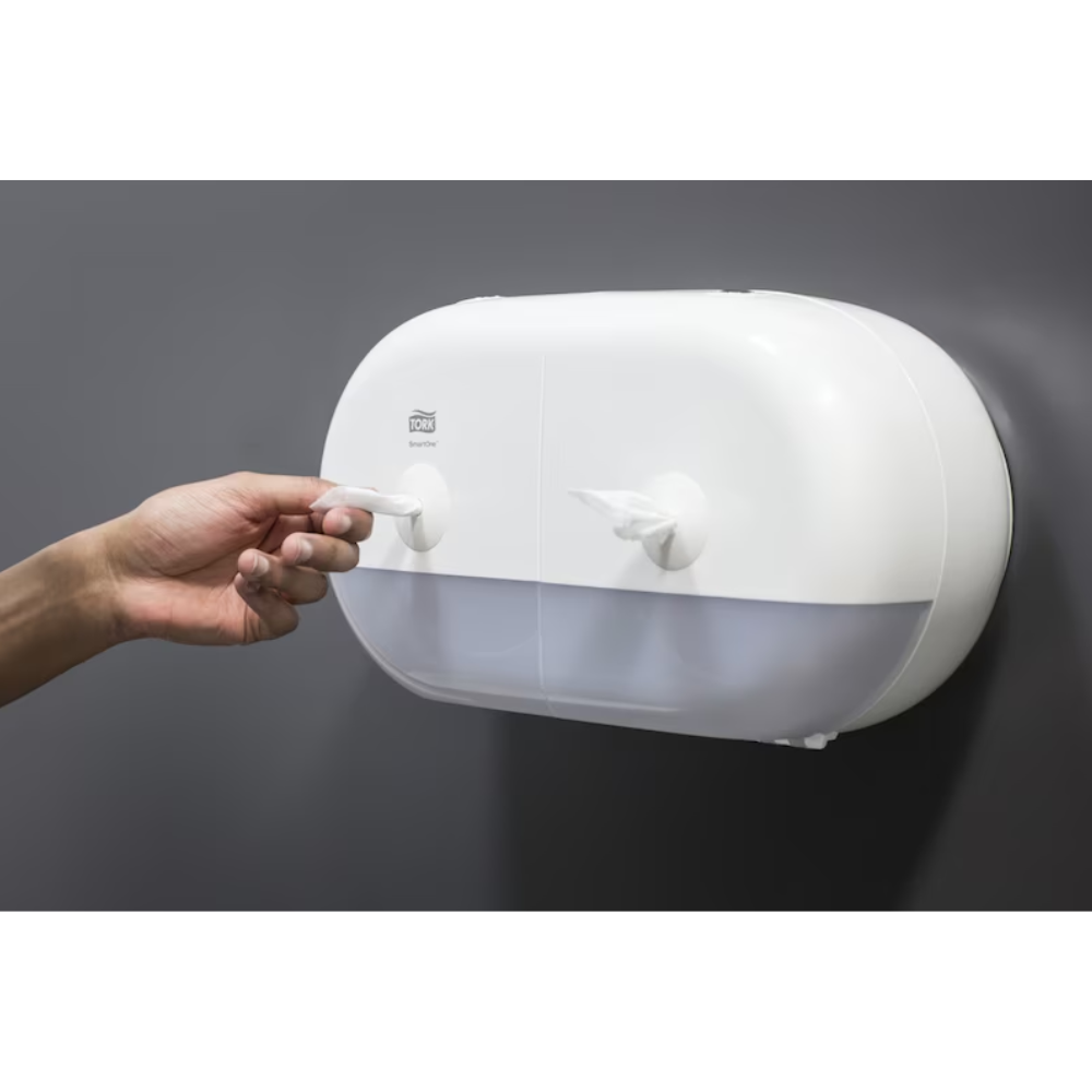 A person pulls a paper towel from an elegant white Tork SmartOne® 682000 Mini Twin Roll Dispenser for toilet paper, which has two compartments and is mounted on a smooth, dark gray wall.