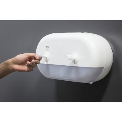 A person pulls a paper towel from an elegant white Tork SmartOne® 682000 Mini Twin Roll Dispenser for toilet paper, which has two compartments and is mounted on a smooth, dark gray wall.