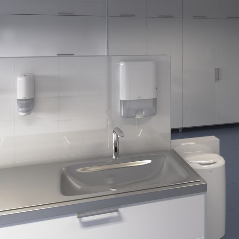Shown is a modern sink in a public restroom with an automatic faucet. Above the sink, a Tork Xpress® 552000 dispenser for Multifold Hand Towels H2 by Essity Professional Hygiene Germany GmbH and a soap dispenser are mounted on a tiled wall. In the background, a row of white cabinets can be seen.