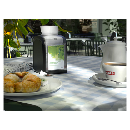 A table in a café, adorned with a blue and white checkered tablecloth, is elegantly set for one person. On the table are a croissant, a cup of coffee in a "Café" cup, a teapot, and a newspaper. The Tork Xpressnap Fit® 272900 table dispenser for napkins by TORK – Essity Professional Hygiene Germany GmbH sits prettily beside it. Sunlight filters through the nearby plants.
