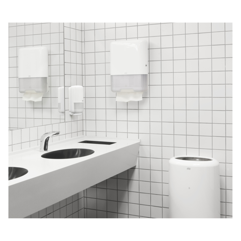 A modern public restroom features a white sink with two faucets, a white tiled wall, and a TORK Tork 100278 extra soft zigzag towels Premium H3 2-ply | Carton (15 packs) dispenser with high-quality 2-ply paper towels. Complemented by a soap dispenser and a trash can, the environment is clean and minimalist.