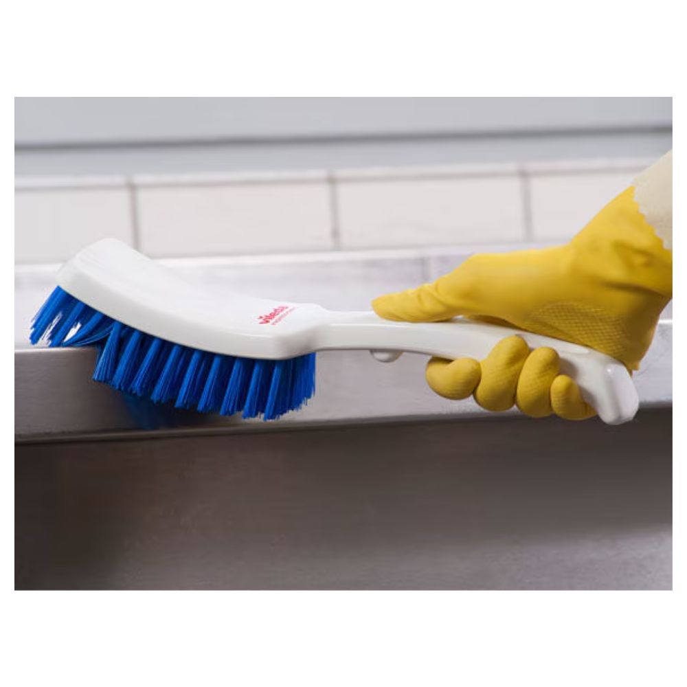 A person with yellow rubber gloves scrubs a gray surface with a Vileda Professional short-handled brush blue – 30 cm by Vileda GmbH, with a tiled wall providing a neutral background.