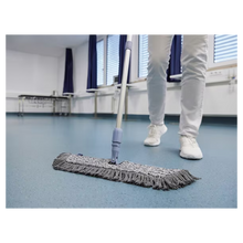 Eine Person in weißen Schuhen und Hosen wischt mit einem Vileda GmbH Professional Swep Duo r-MicroCombi Mop - 50 cm einen blauen Boden in einem Raum mit geschlossenen Jalousien und blauen Vorhängen. In der Nähe stehen ein Klapptisch und unter den Fenstern ein Heizkörper.