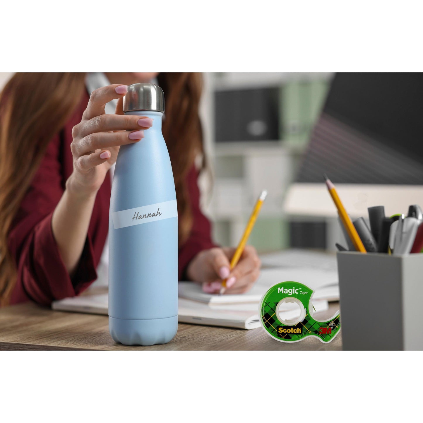 A person with a blue bottle labeled "Hannah" sits at a desk with notebooks, pens, and the Scotch® Magic™ Invisible Tape 8-1915R3/2 promotional pack from 3M - ideal for crafting. In the background are office shelves and a blurred work area.
