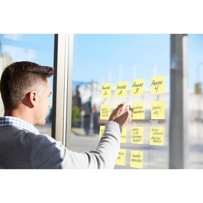 A man uses the 3M Deutschland GmbH Post-it® Z-Notes Dispenser, PRO (black) and yellow Post-it® Super Sticky Z-Notes (76x76 mm, 90 sheets/block), to organize project tasks on a glass window with sunlight and a street outside.