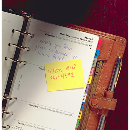 An open leather planner shows notes for a lunch at 1 PM with John and a press conference at 5 PM at the Hilton Hotel. On a Post-it® Notes Cube 636-B, Yellow, 76 mm x 76 mm from 3M Germany GmbH, it says "Hilton Hotel 761-4392". A pen lies next to it.
