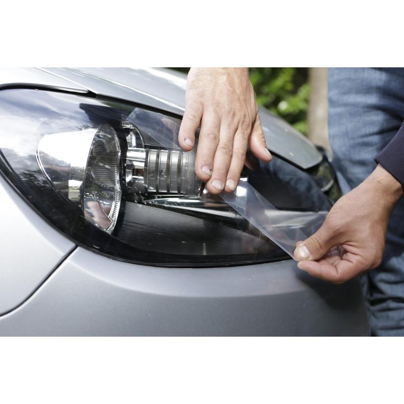 A person uses 3M Deutschland GmbH Scotch® Extremium™ INVISIBLE High-Performance Tape (20 m x 48 mm) to attach a protective clear film to a silver car headlight, smoothing the film by hand over the surface.