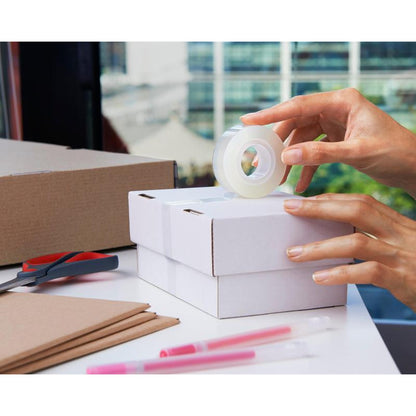 A person seals a small white box with Scotch® Crystal Tape, transparent, value pack (8 rolls, 19 mm x 33 m) from 3M Deutschland GmbH. Scissors, cardboard pieces, boxes, and pink pens are on the table near a window.