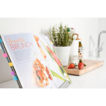 An open cookbook with the Post-it® Index Tabs Mini from 3M Deutschland GmbH marks a brunch recipe. In the background, strawberries, a pot with herbs, and kitchen utensils are arranged on a wooden board on a white countertop.