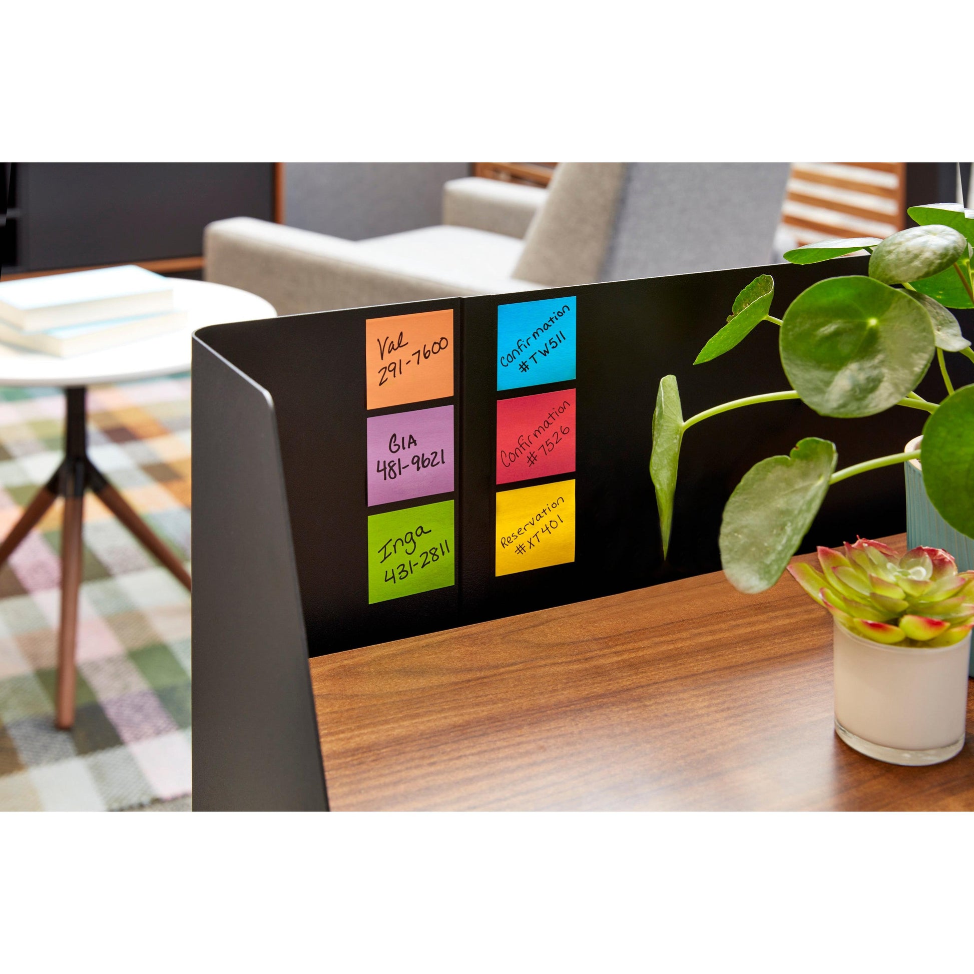 Ein schwarzer Schreibtischteiler mit 3M Post-it® Super Sticky Z-Notes in verschiedenen Farben (76x76mm, 14+2 Promo-Pack) mit handschriftlichen Informationen steht neben einer grünen Pflanze auf einem hölzernen Bürotisch.