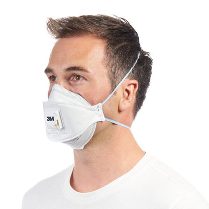 A man with short brown hair, in a plain white shirt, wears a 3M™ Aura™ Comfort Cool Flow™ Particulate Respirator 9322+ FFP2 with valve from 3M Deutschland GmbH, secured with elastic bands and slightly facing left.