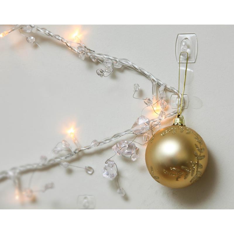 A golden Christmas ornament hangs on a Command™ Mini Hook, transparent, with transparent strips, 17006CLR from 3M Deutschland GmbH, next to illuminated white lights and clear crystals on a white wall.