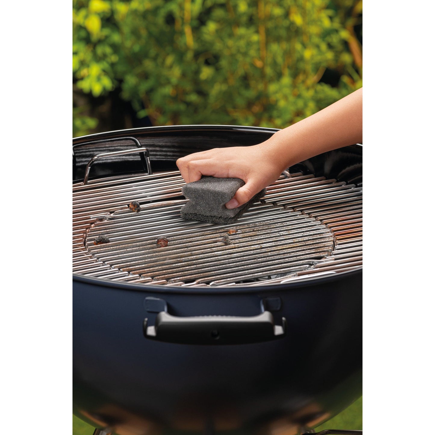 A hand cleans the metal grill of a round charcoal grill with a Scotch-Brite® Grill Handle Sponge (1 piece per package) from 3M Deutschland GmbH. Green foliage is blurred in the background.