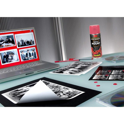 A workspace with a laptop with photo editing software, black and white prints, a can of 3M™ Scotch® PhotoMount™ adhesive spray (400 ml, 1 can, 3M Deutschland GmbH), an empty sheet for mounting, a transparent CD case, and red adhesive dots on a glass table.