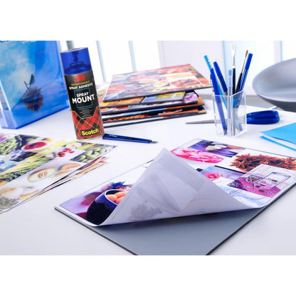 A creative workspace by the bright window with glossy pictures, brushes, pens, scissors, folders, and the 3M™ Scotch® SprayMount™ Adhesive Spray (400 ml, 1 can/package) from 3M Deutschland GmbH - ideal for photo collages and crafts.
