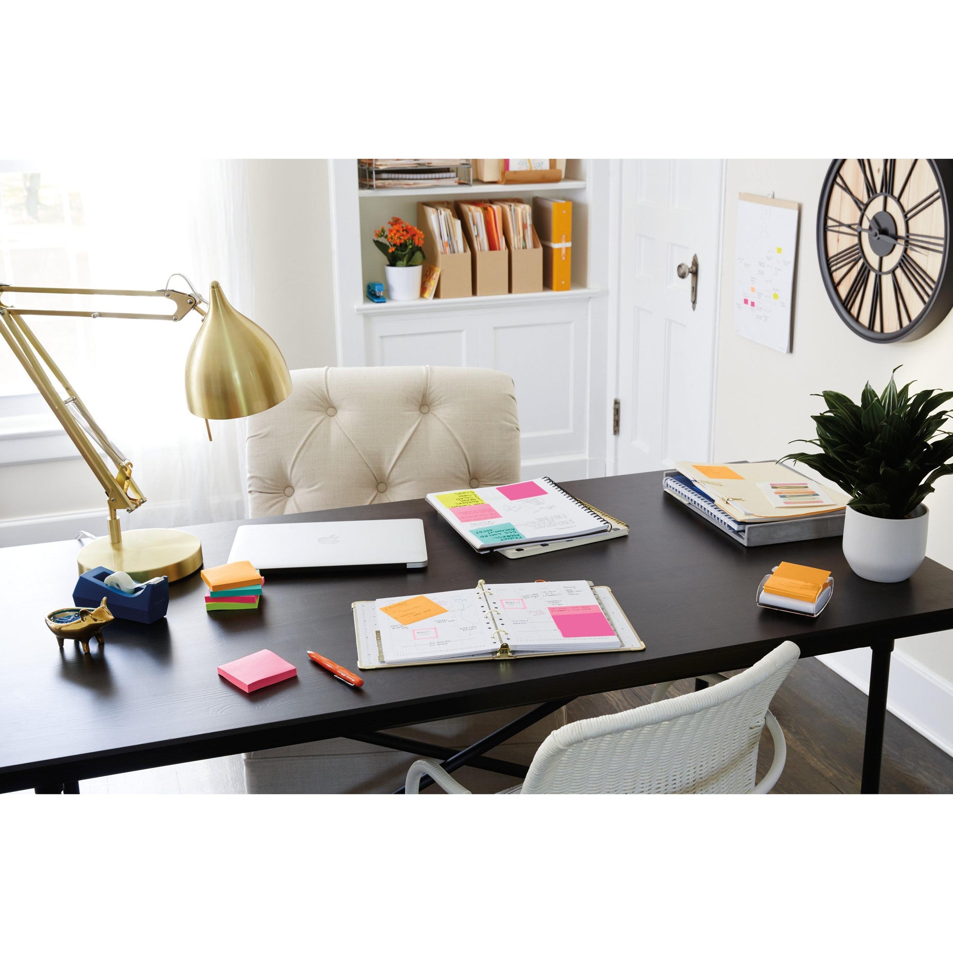 Ein aufgeräumtes Heimbüro mit schwarzem Schreibtisch zeigt einen Laptop, Planer, ein 3M Deutschland GmbH Post-it® Super Sticky Notes 654-6SS-PPNK Paket (rosa, 76x76mm), Lampe, Stifthalter, Pflanze und Dekoration. Im Hintergrund befinden sich Regale, Akten und eine große Wanduhr.
