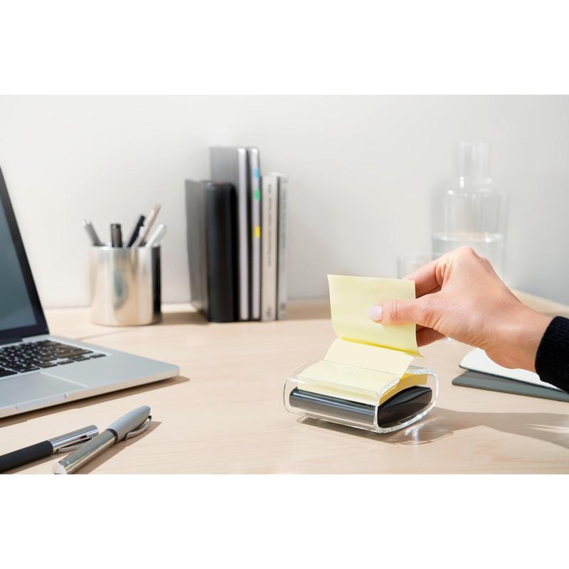 A hand pulls a yellow Post-it® Super Sticky Z-Note (76x76 mm) from the black PRO Post-it® Z-Notes Dispenser from 3M Deutschland GmbH on a tidy desk with a laptop, pens, books, glass carafe, and cup neatly in the background.
