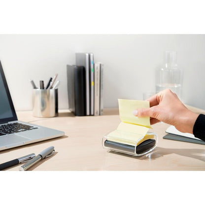 A hand pulls a yellow Post-it® Super Sticky Z-Note (76x76 mm) from the black PRO Post-it® Z-Notes Dispenser from 3M Deutschland GmbH on a tidy desk with a laptop, pens, books, glass carafe, and cup neatly in the background.