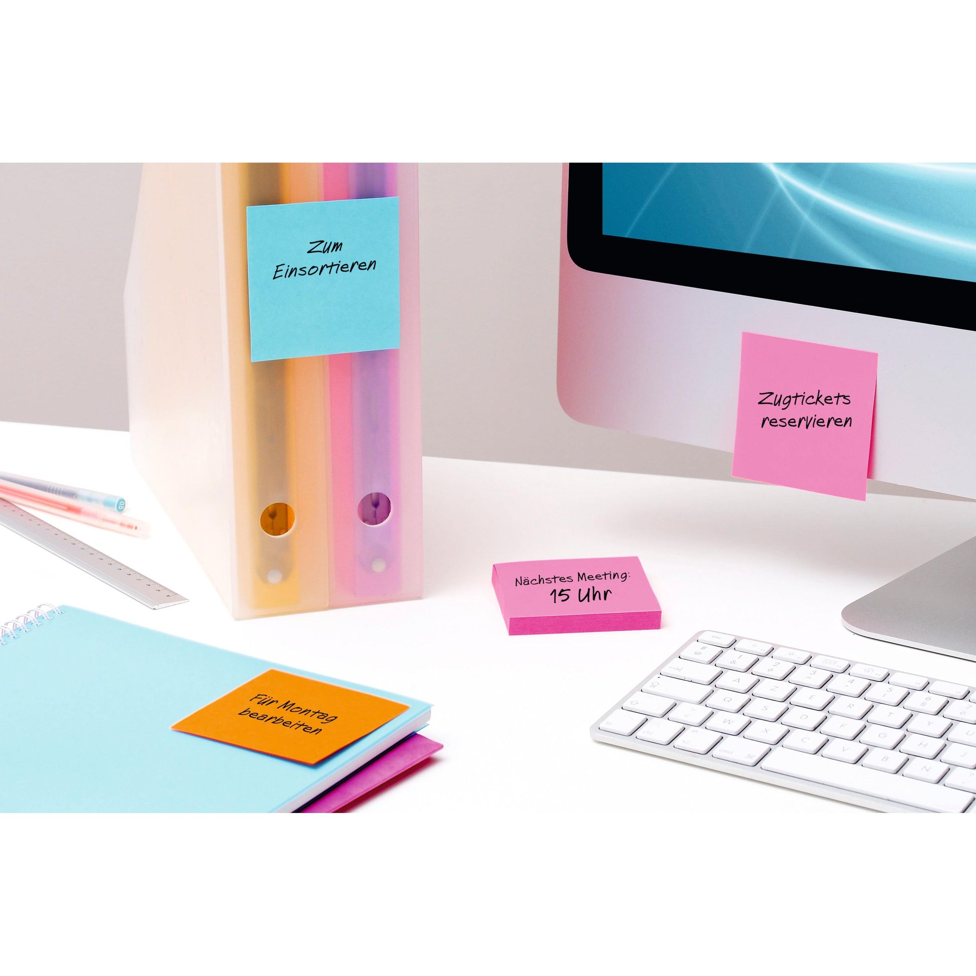 Ein aufgeräumter Schreibtisch mit Computer, Tastatur, Linealen, Notizbüchern, Ordnern und 3M Deutschland GmbH Post-It® Super Sticky Notes (4 Blöcke/Packung), bunt und deutsch beschriftet mit Erinnerungen wie Meetings und Zugtickets.